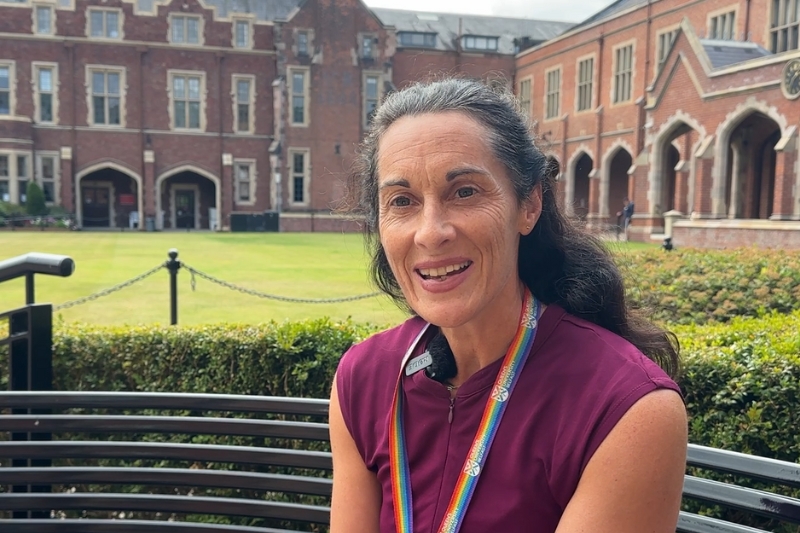Dark haired lady sitting on a black bench in the quad of QUB with a grassy area as a backdrop